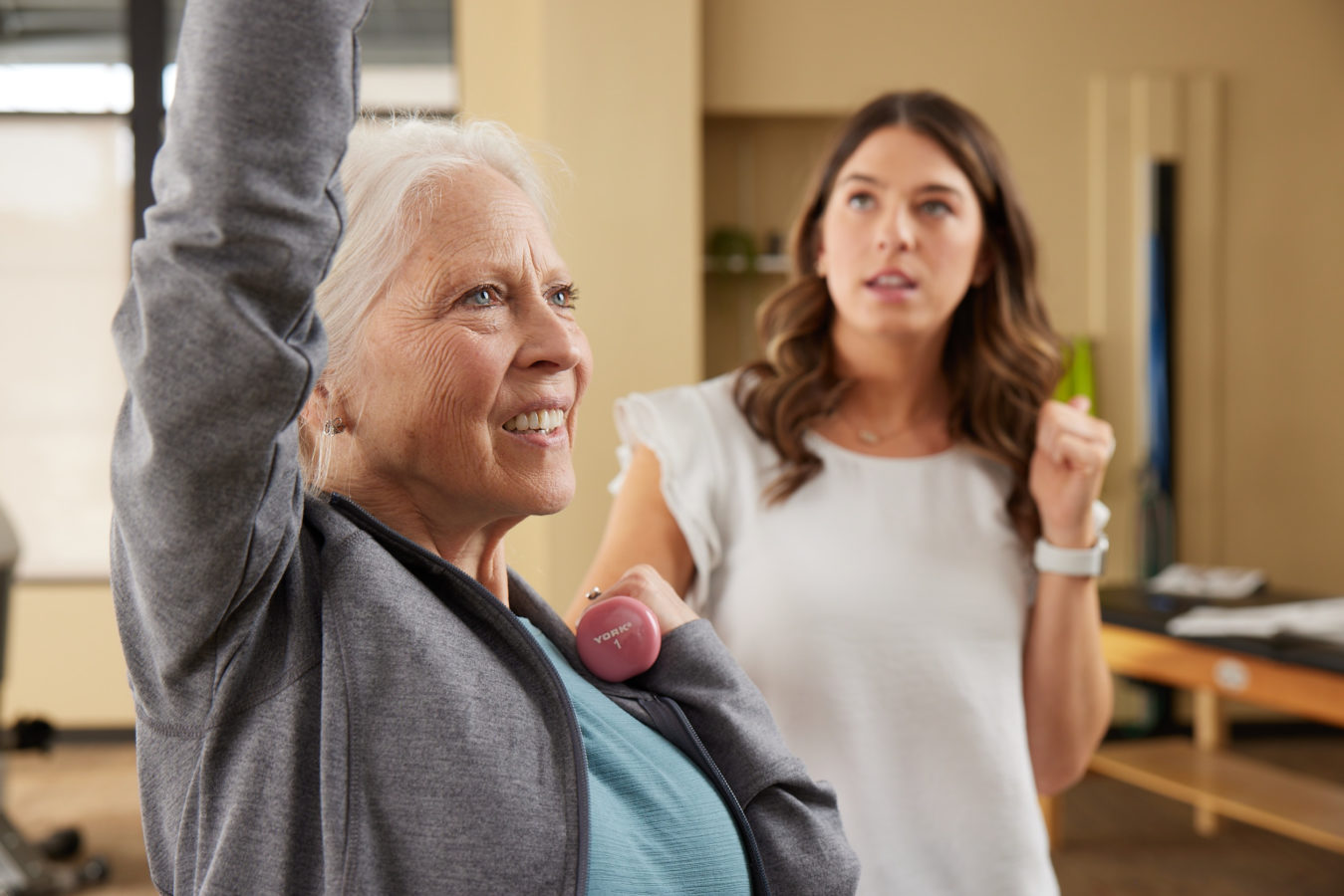 older woman with white hair smiling as she lifts small weights with female physical therapist