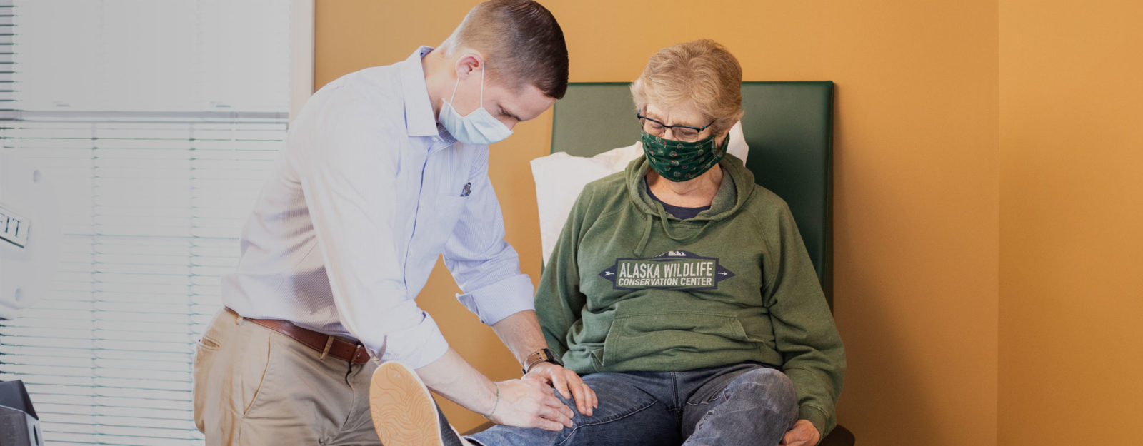 young male physical therapist in a mask examining older female patient's knee